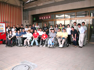 アイ・コラボレーション事務所の前の広い駐車場に、サポーターや、車いすの人、杖をついた人等、数十人のスタッフが並んでいる集合写真