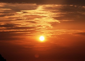 オレンジ色の夕焼け空に、黄色がかった丸い太陽があり、オレンジがかった穏やかな海面が太陽光を反射しています