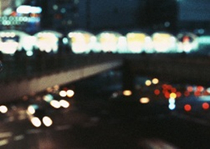 夜のJR○○駅前交差点。十字に交差した歩道橋の下に、同じく十字に交差した道路があり、交通量が多めです。車の白いライトや赤いブレーキランプ、ビルの看板の明かりや街灯等、沢山の明かりが暗闇に光っています